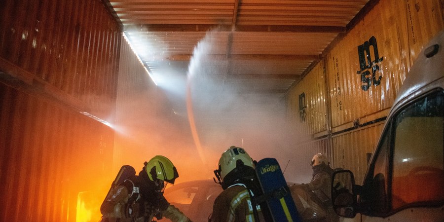 Brandweermensen in een tunnel van containers die een brandend voertuig blussen